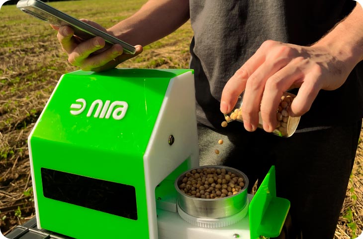 Homem no campo segurando celular enquanto despeja soja em uma máquina verde com uma logo escrito Nira